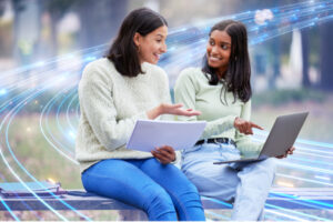 Young ladies sitting and pointing at laptop while holding a notebook up.