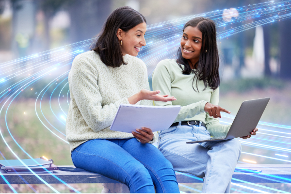 Young ladies sitting and pointing at laptop while holding a notebook up.