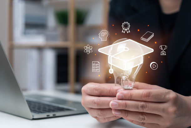 Person holding a lightbulb with a glowing graduation cap icon and educational symbols, sitting at a desk with a laptop.