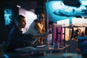 Two colleagues collaborating at a computer in a futuristic, tech-focused workspace with neon lighting and digital displays in the background.