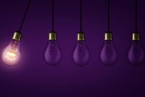 Five lightbulbs with one lit and purple dark background.