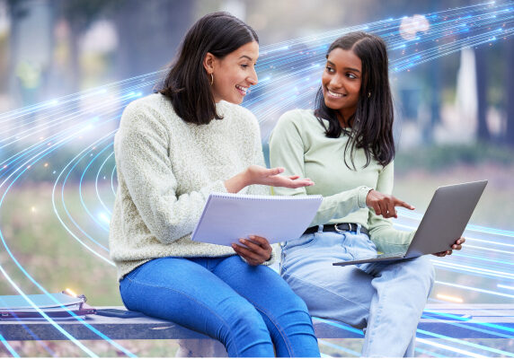 Young ladies sitting and pointing at laptop while holding a notebook up.