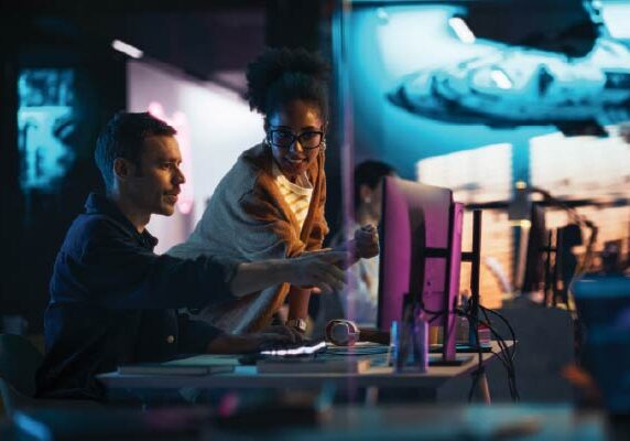 Two colleagues collaborating at a computer in a futuristic, tech-focused workspace with neon lighting and digital displays in the background.