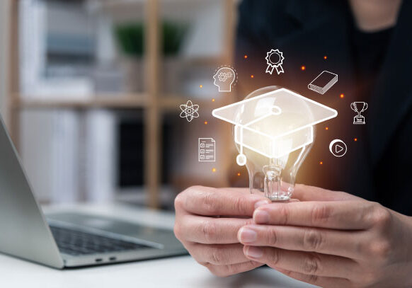 Person holding a lightbulb with a glowing graduation cap icon and educational symbols, sitting at a desk with a laptop.